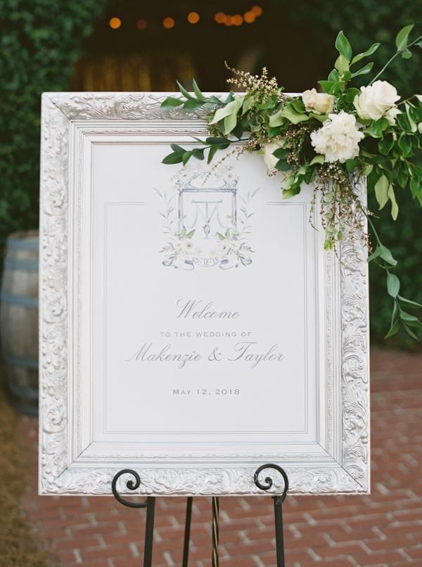 a sign with flowers and greenery on it sitting in front of a brick walkway