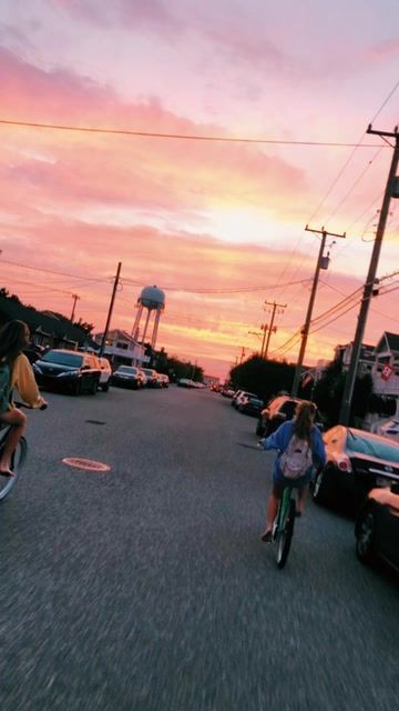 two people riding bikes down a street at sunset with cars parked in the parking lot behind them