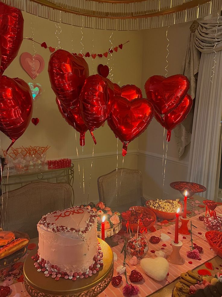 a table topped with lots of red heart shaped balloons and cake covered in white frosting