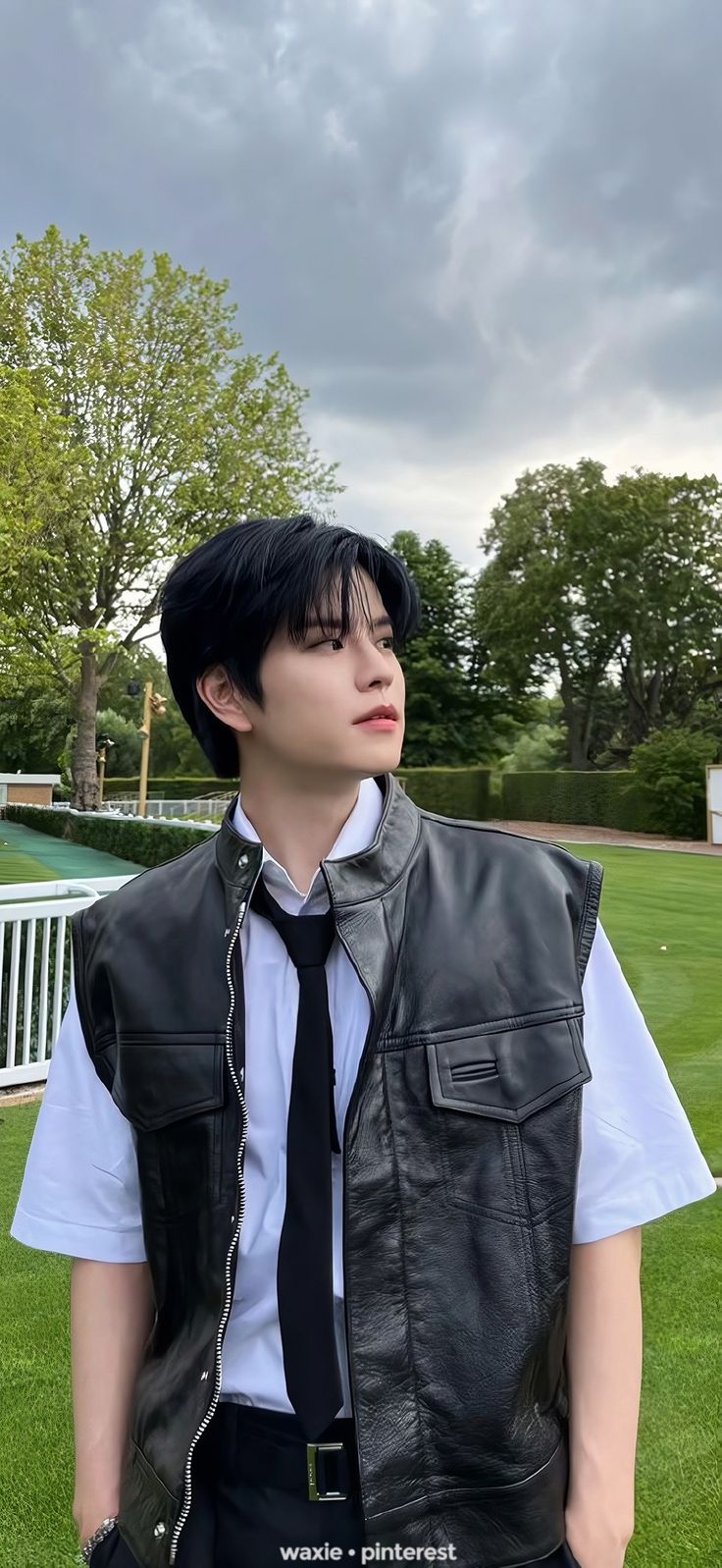 a young man wearing a black leather vest and tie standing on grass in front of a white fence