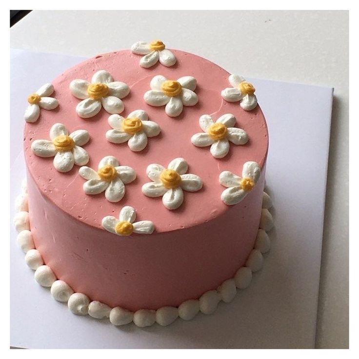 a pink cake with white and yellow flowers on it sitting on top of a piece of paper