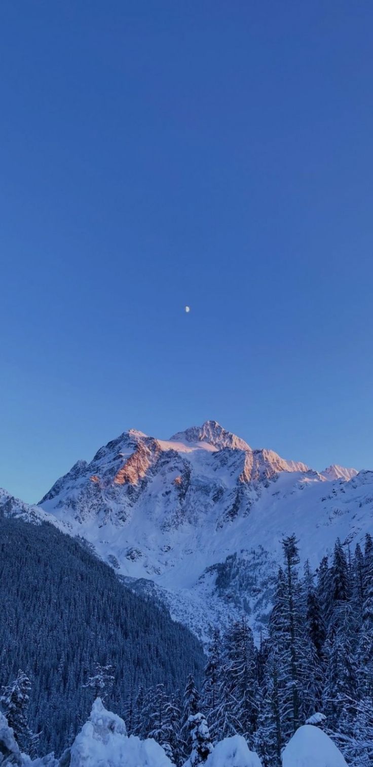 the mountains are covered in snow and there is a half moon rising above them on a clear day