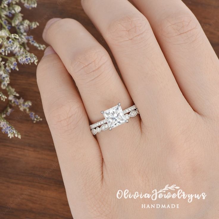 a woman's hand with a diamond ring on top of her finger and flowers in the background