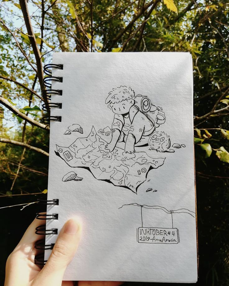 a hand holding a notebook with a drawing on it in front of trees and bushes