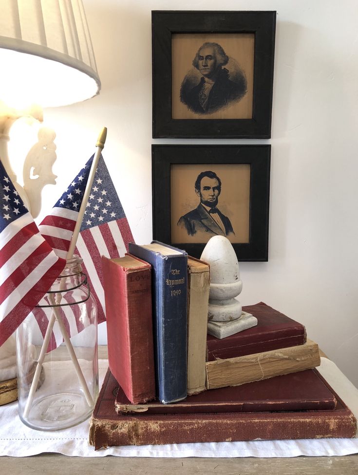 an american flag and books on a table with two framed pictures in the back ground