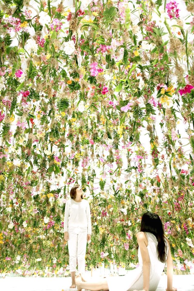 two people sitting on the ground in front of a wall with flowers hanging from it