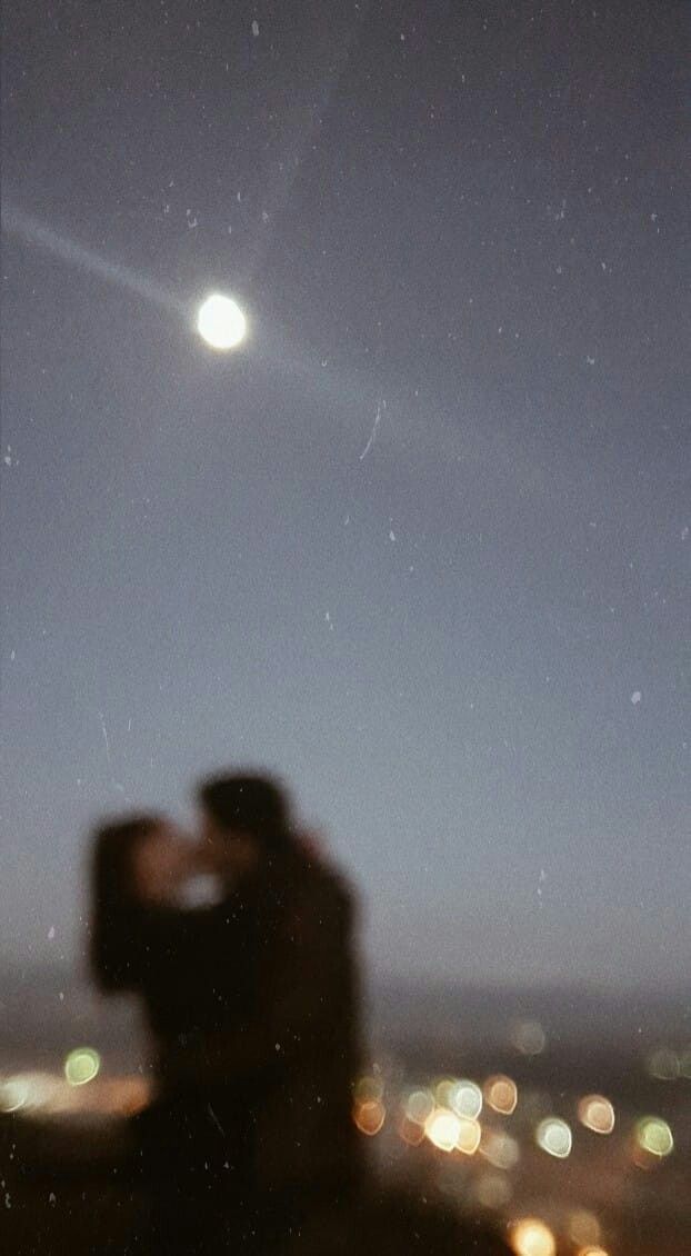 a man and woman are kissing in front of the city lights at night, while the moon shines brightly above them