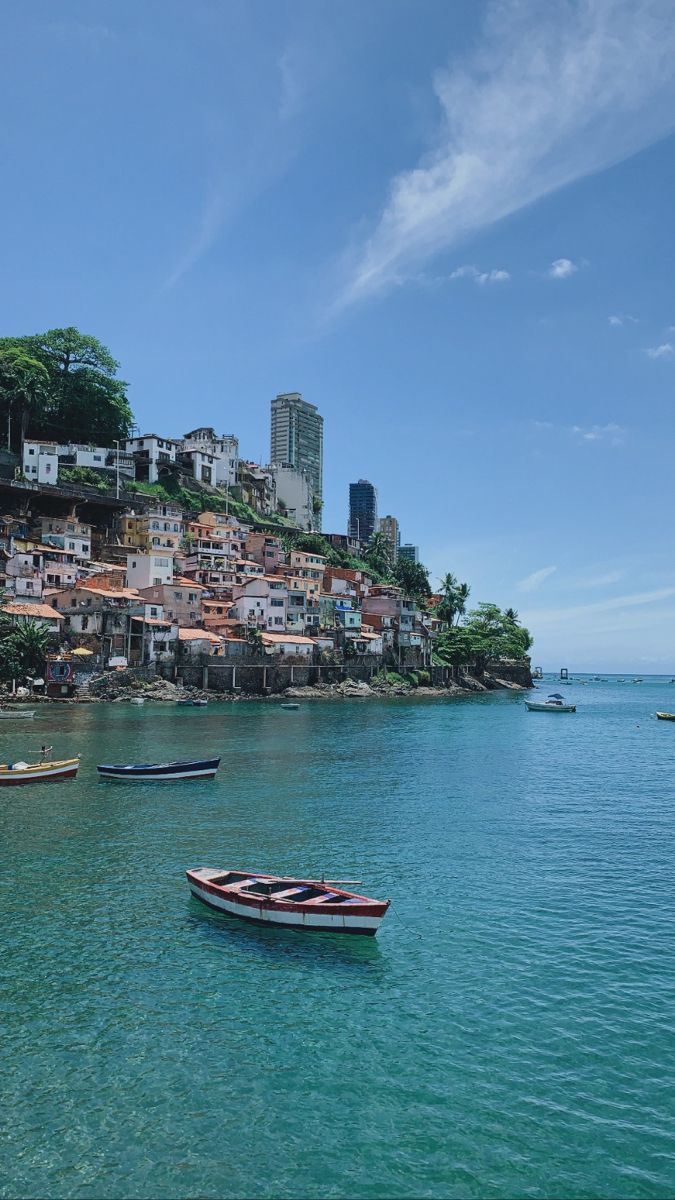 several small boats floating on the water in front of a hill with houses and buildings