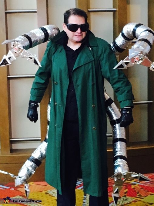 a man in green coat and sunglasses standing next to giant metal arms with claws on each arm