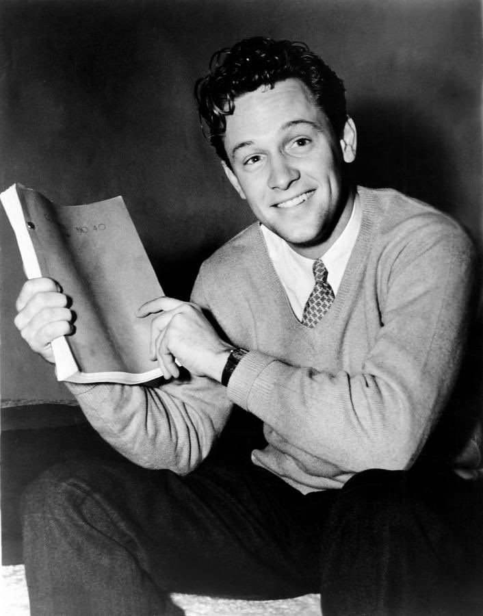 a man in a sweater and tie is holding a book while sitting on the floor