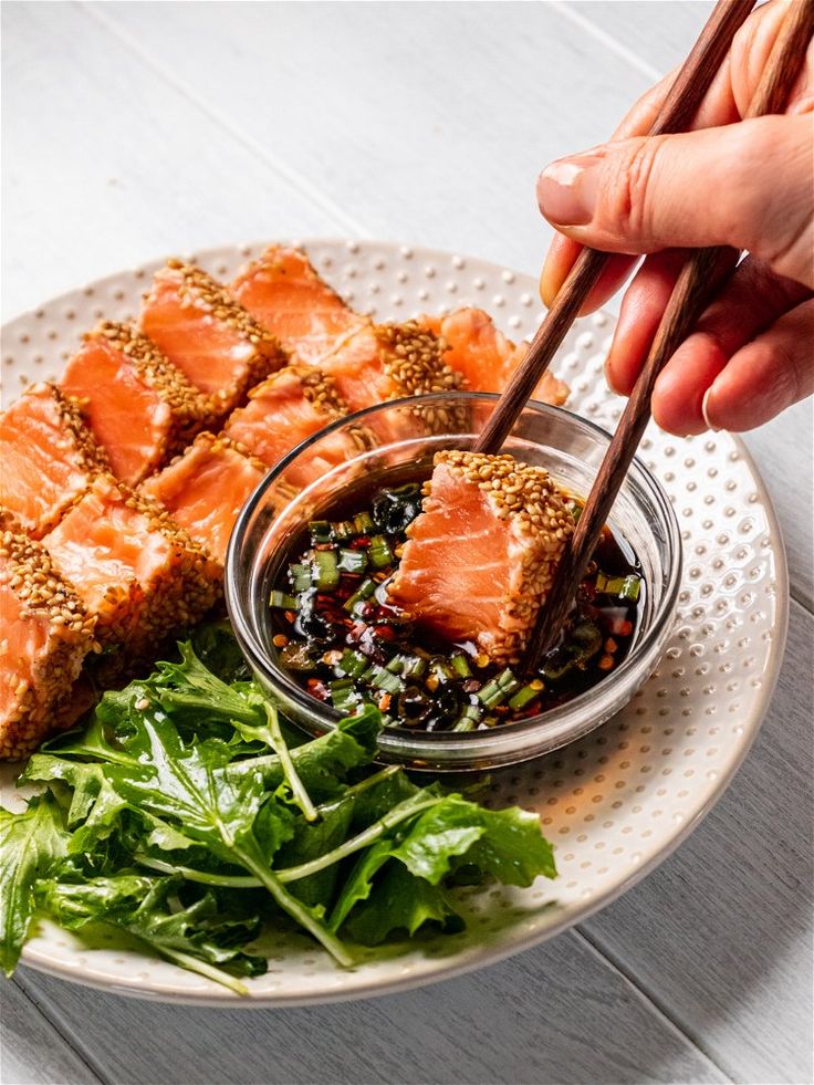 a plate with salmon, greens and sesame seeds on it is being held by a pair of chopsticks