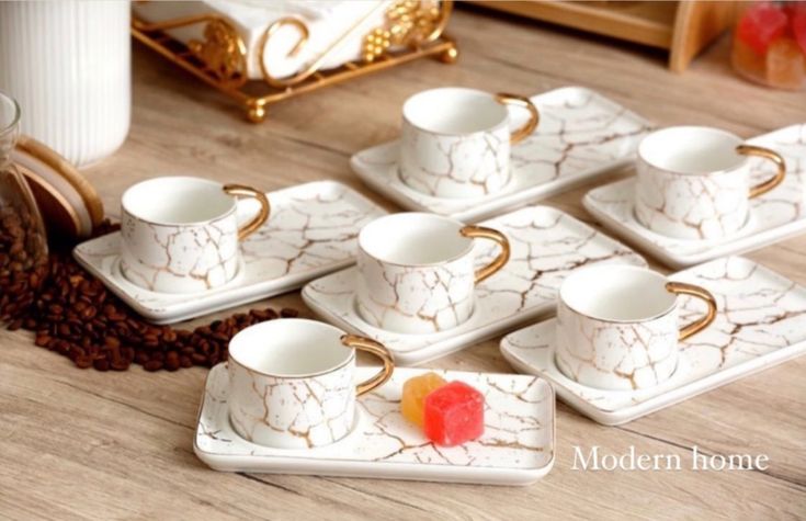 six white and gold coffee cups with saucers on a wooden table next to a red piece of candy