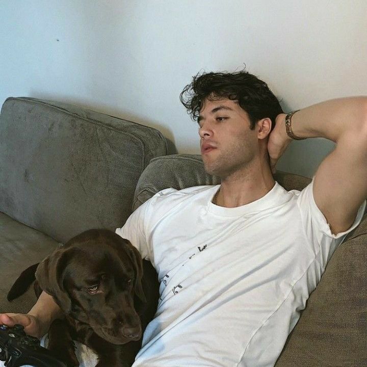 a man laying on top of a couch next to a brown dog and holding a game controller