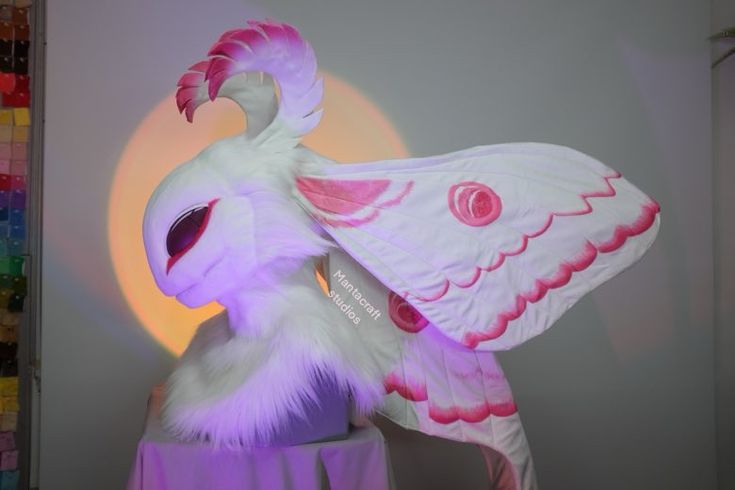 a white and pink stuffed animal with wings on it's head standing in front of a wall