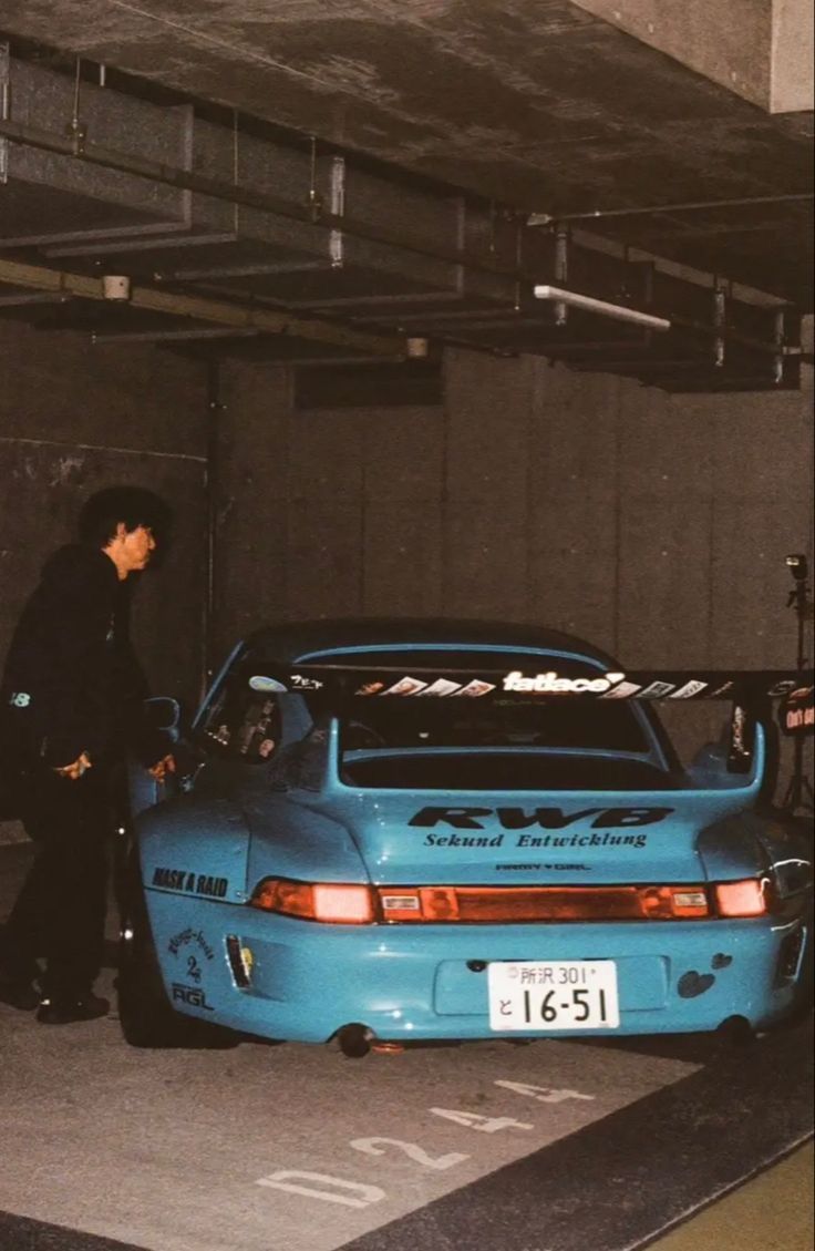 a man standing next to a blue car in a parking garage with the hood up