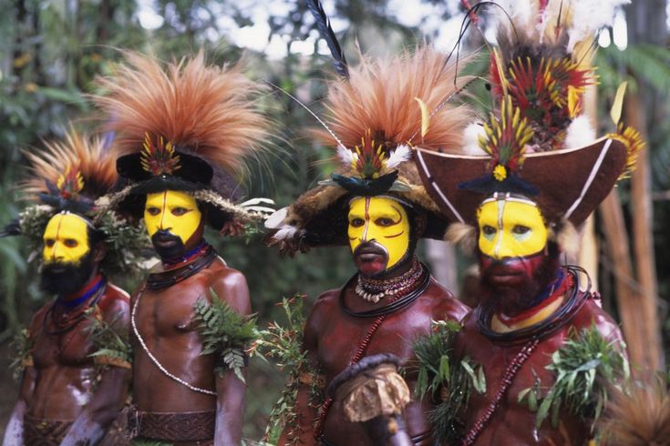 three men with painted faces and headdresses are standing in front of trees