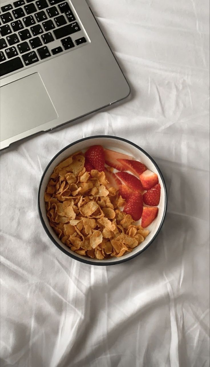 a bowl of cereal and strawberries next to a laptop computer on a white sheet