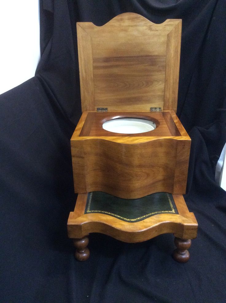 a wooden toilet seat with an open lid sitting on a black cloth covered floor in front of a white wall