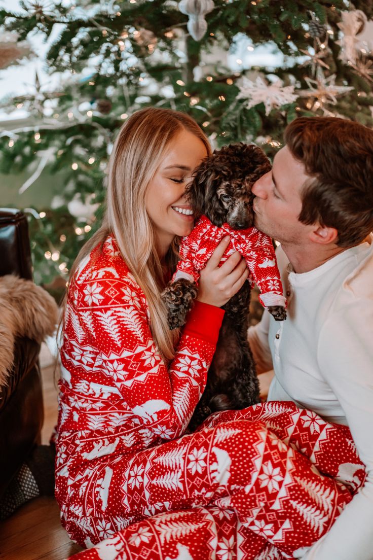 a man and woman kissing while holding a dog in front of a christmas tree