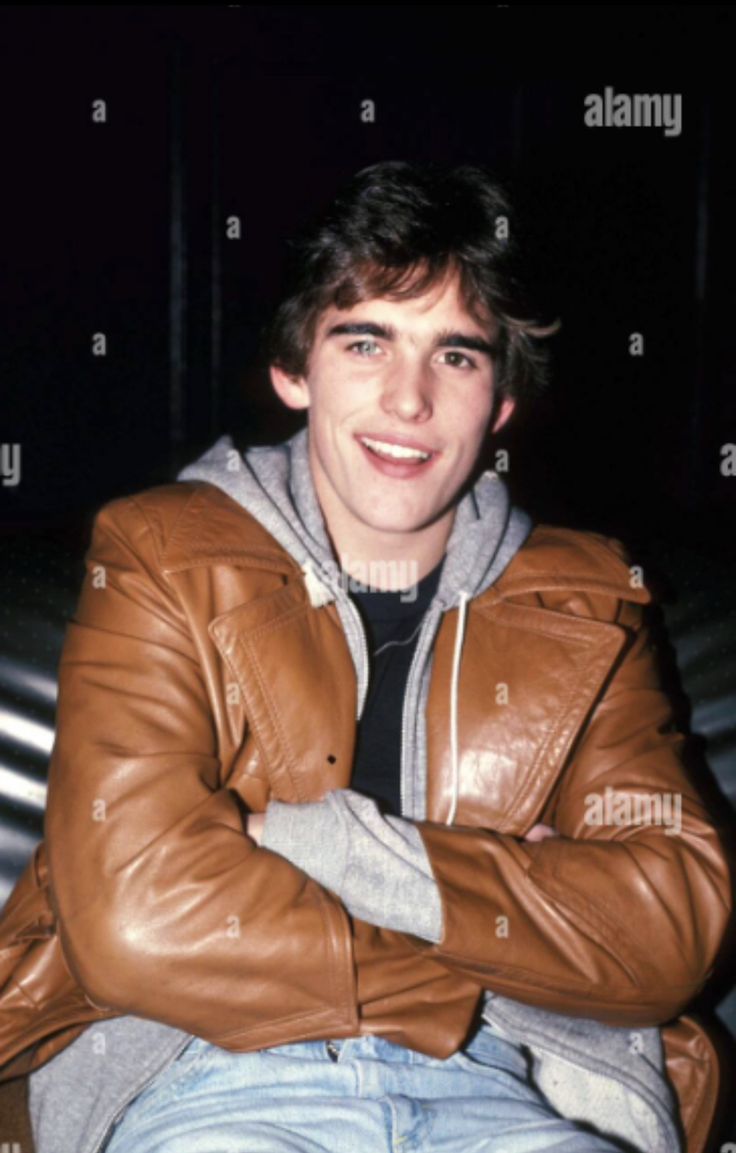 a young man wearing a brown leather jacket and jeans sitting on a bench with his arms crossed - stock image