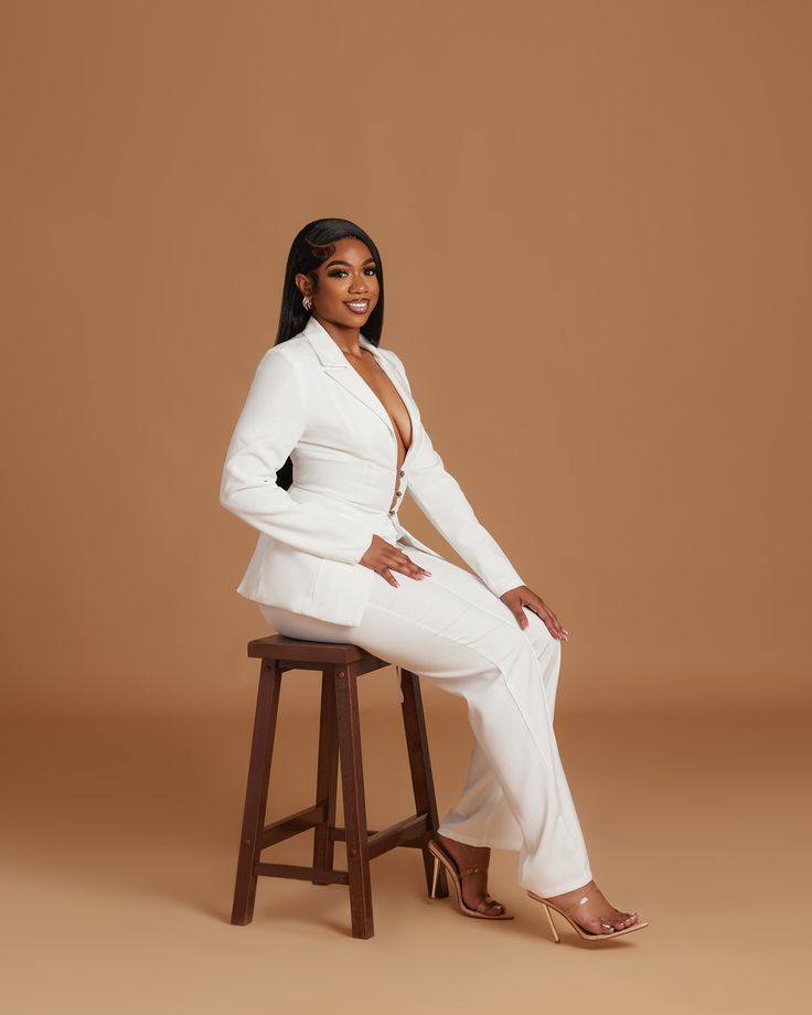 a woman sitting on top of a wooden stool wearing a white suit and high heels