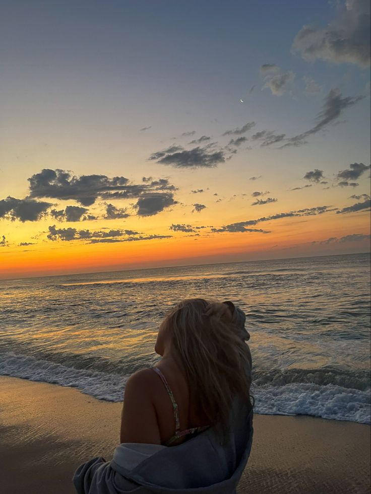 a woman sitting on top of a sandy beach next to the ocean at sun set