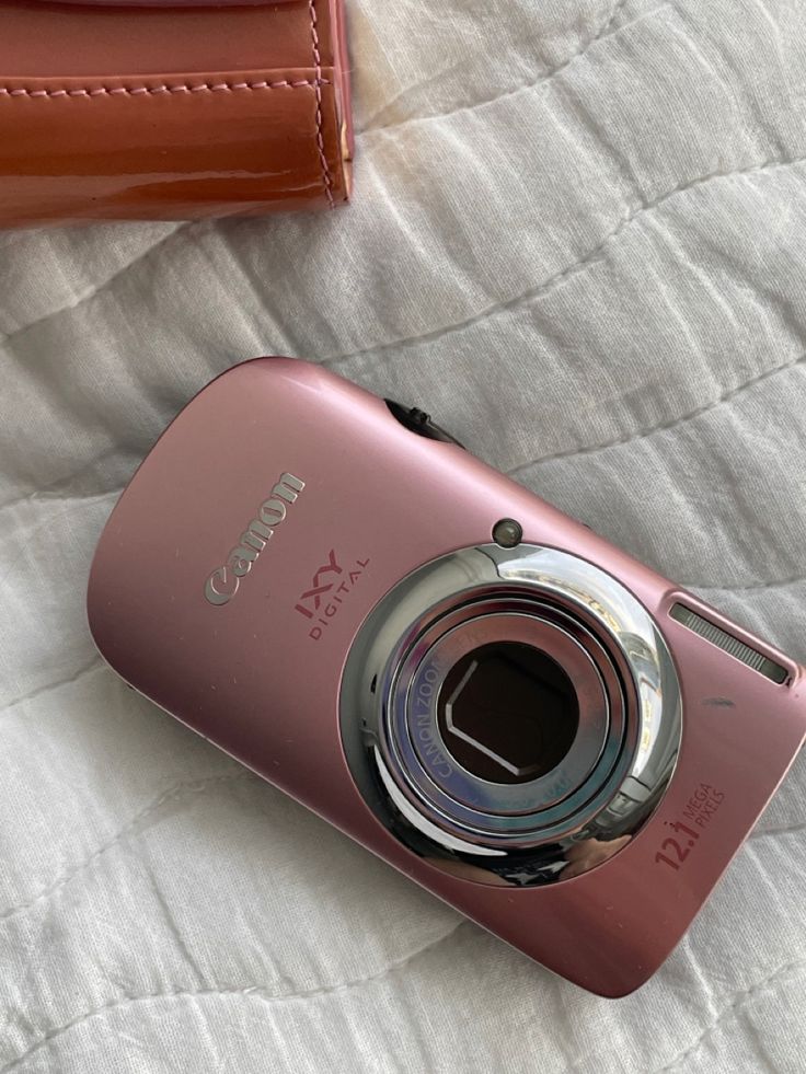 a pink camera sitting on top of a bed next to a brown case and wallet