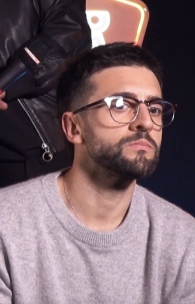 a man with glasses sitting in front of a person wearing a black leather jacket and grey sweater