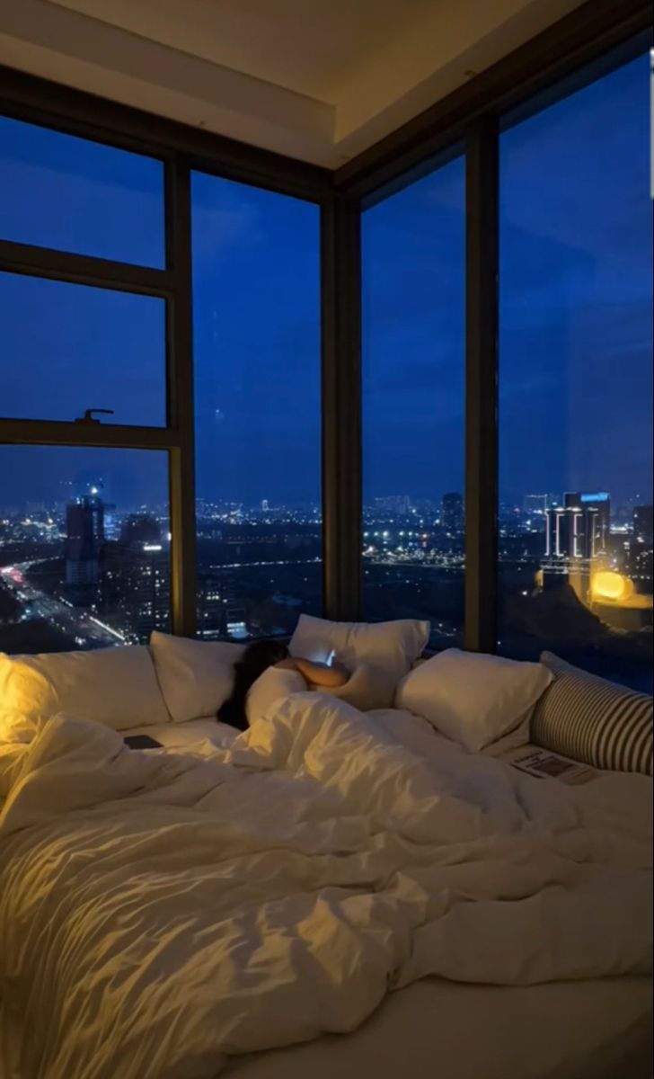 a woman laying on top of a bed in a bedroom next to large windows at night