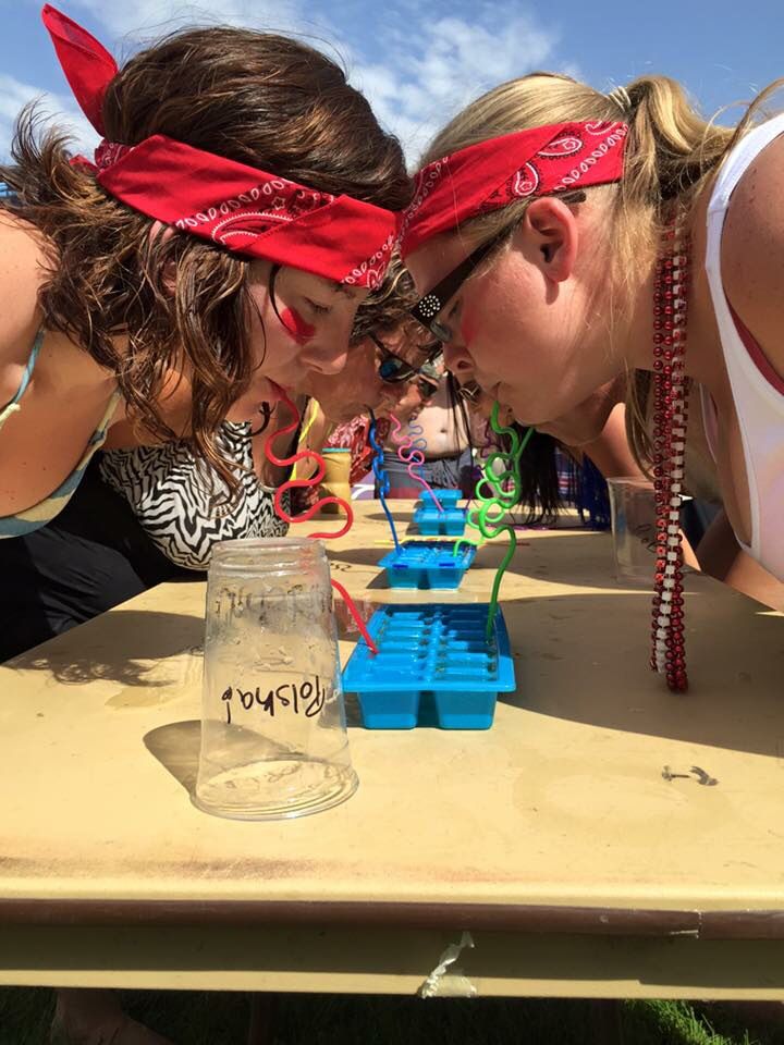 two girls blowing out candles on a table at an outdoor event with other people in the background