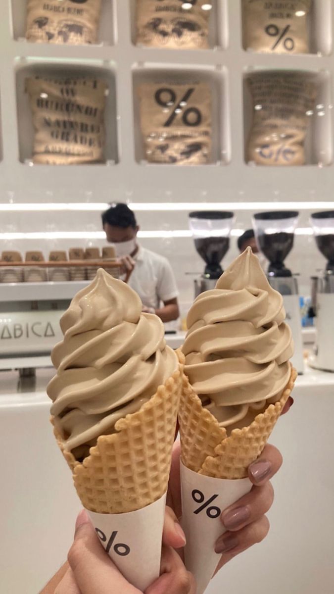 two ice cream cones are being held up in front of a counter with people working behind them