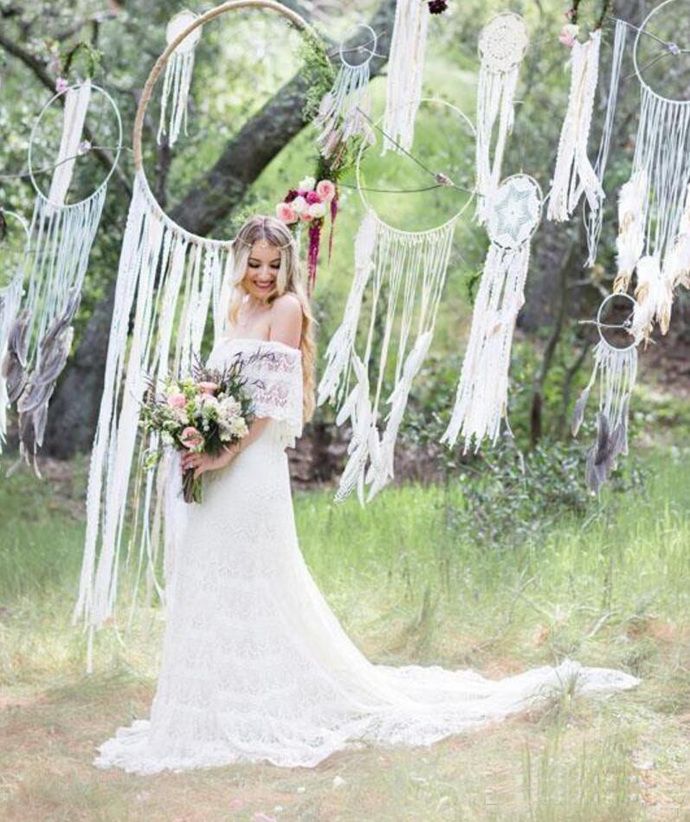 a woman in a wedding dress holding a bouquet and standing under an outdoor dream catcher