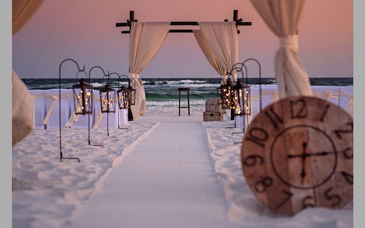 an outdoor wedding set up with white drapes and lights on the beach at sunset