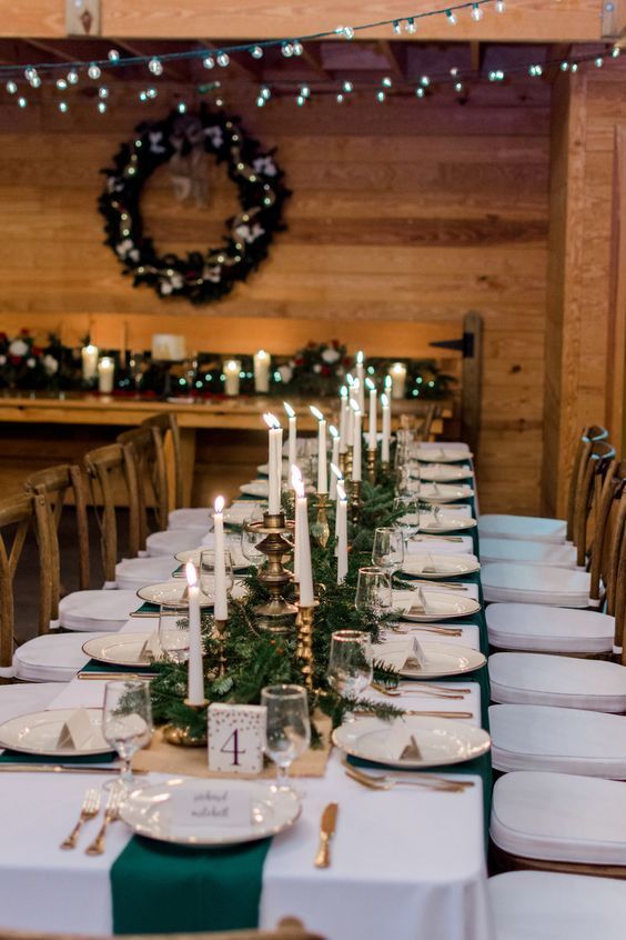 a long table is set with white plates and silverware, candles and greenery