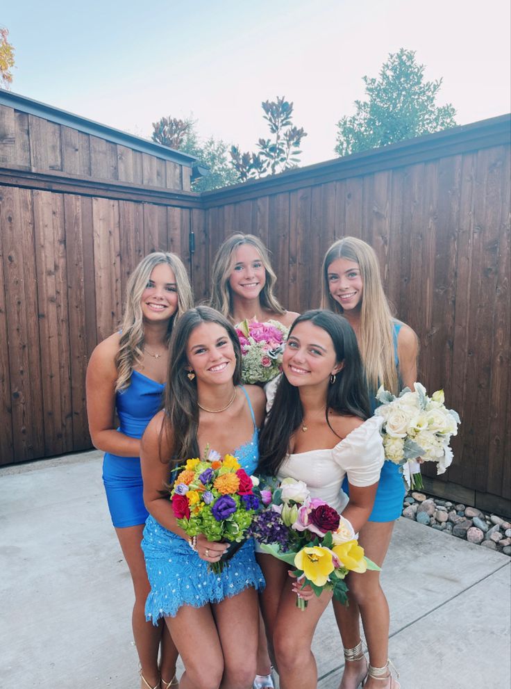 a group of women standing next to each other in front of a wooden fence holding bouquets