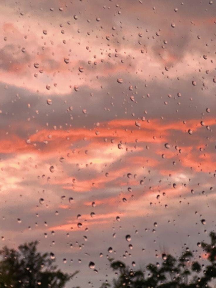 rain drops on the window as the sun sets in the sky behind trees and clouds