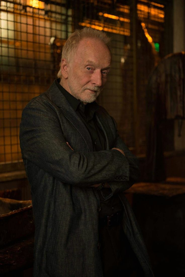 an older man with white hair wearing a black coat and standing in front of a window