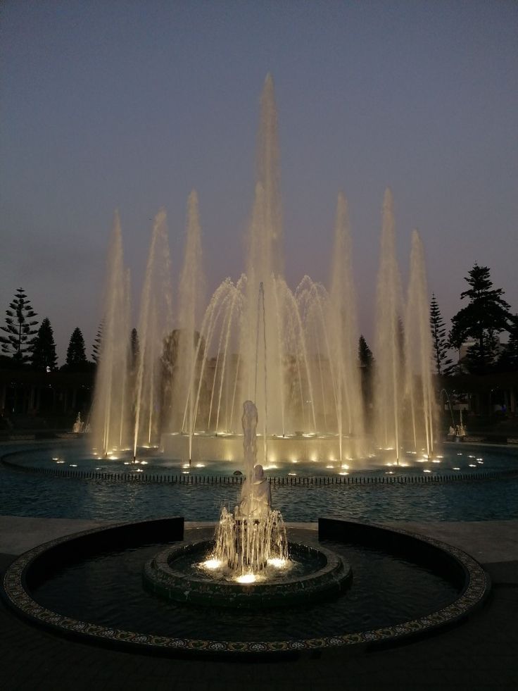 a man standing in front of a fountain at night with his arms up to the sky