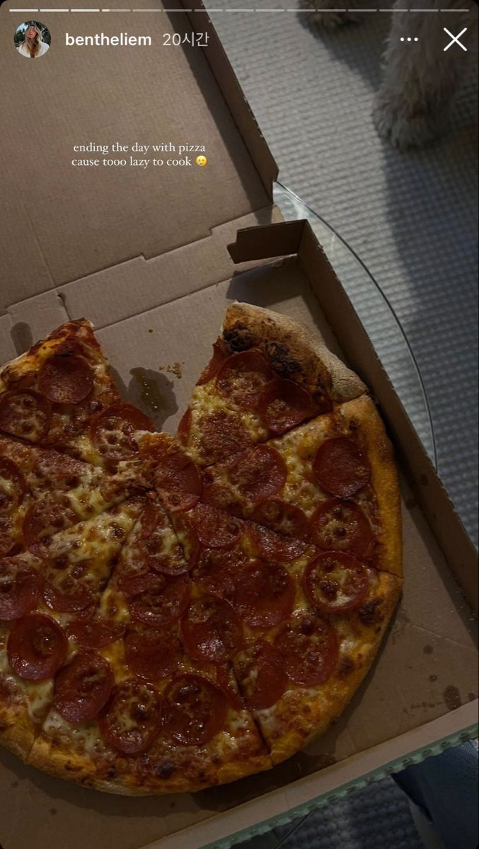 a pepperoni pizza sitting in a box on the floor next to a dog's paw