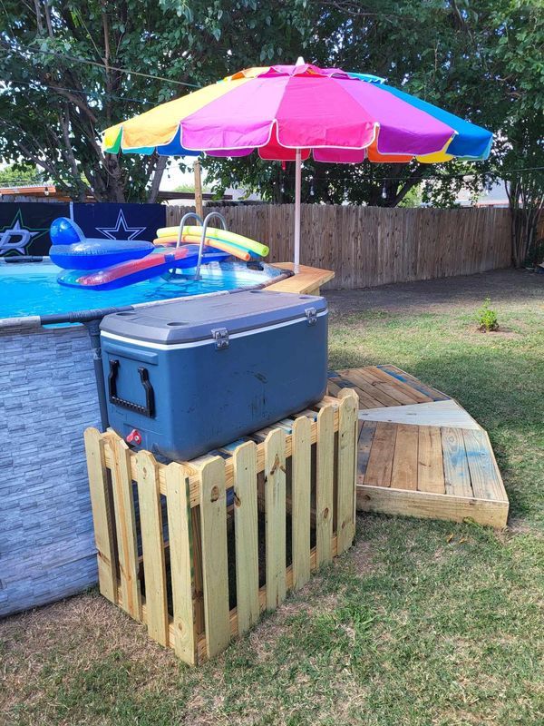 a cooler sitting under an umbrella next to a swimming pool in a backyard with wooden pallets