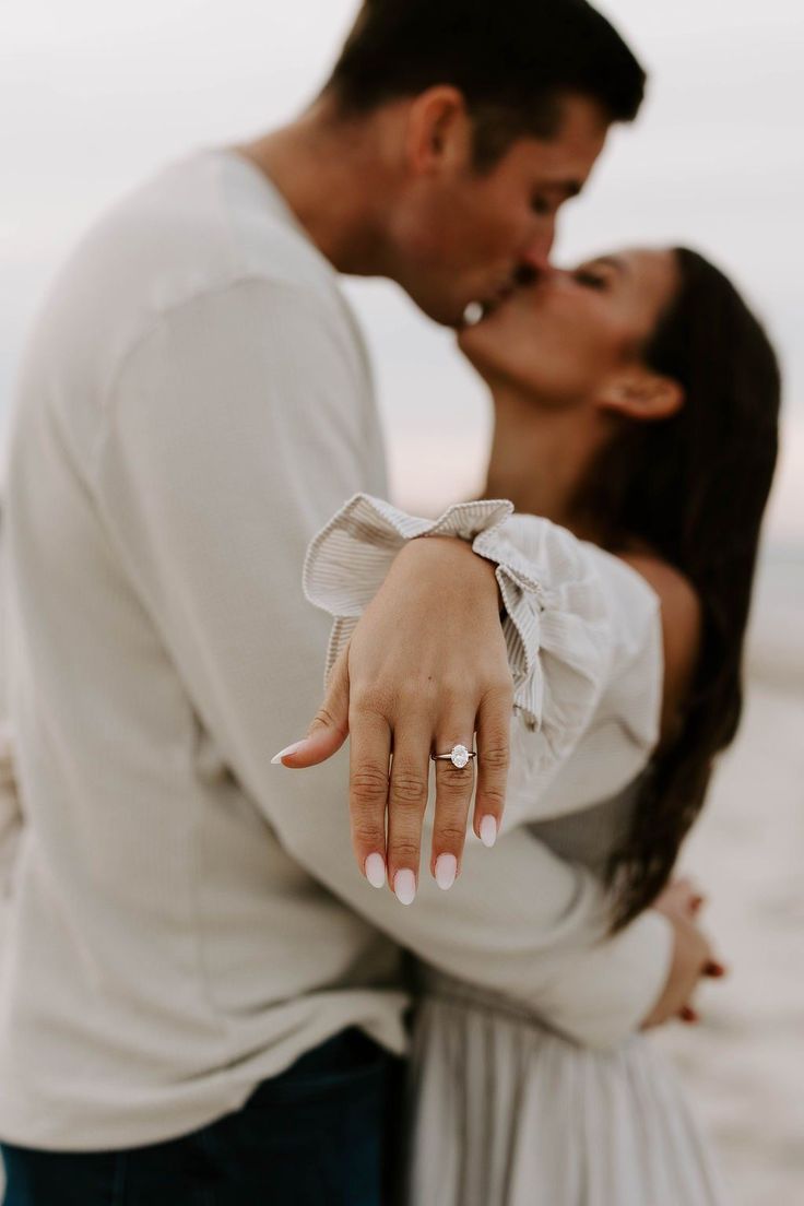 an engaged couple kissing on the beach with their engagement ring in front of him and her