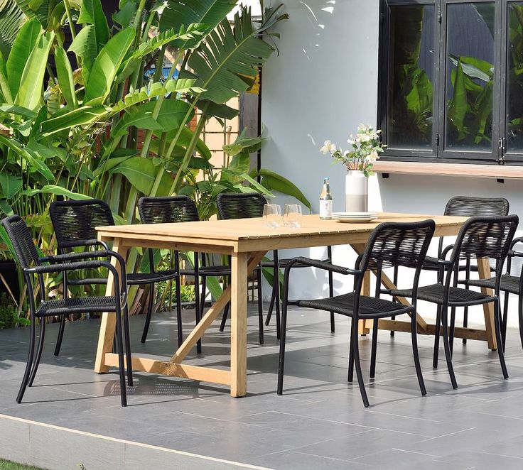 a wooden table with black chairs and plants in front of it on a patio next to a building