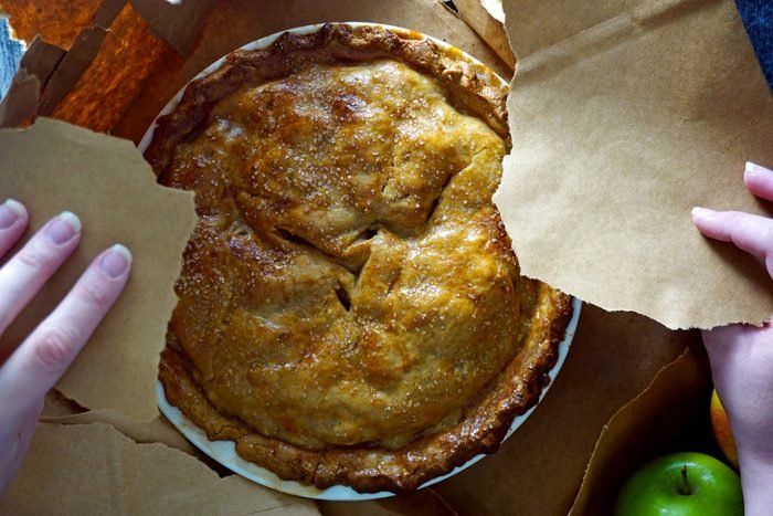 two hands reaching for an apple pie on top of some brown paper and green apples