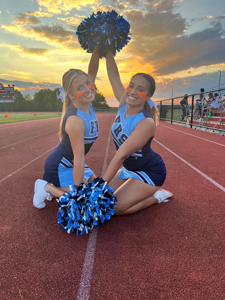 two cheerleaders kneeling on the sidelines with their pom poms