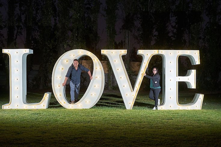 two people standing in front of the word love lit up with light bulbs at night