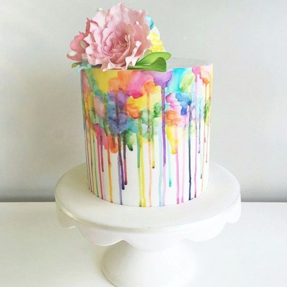 a multicolored cake decorated with flowers and dripping icing on a white pedestal