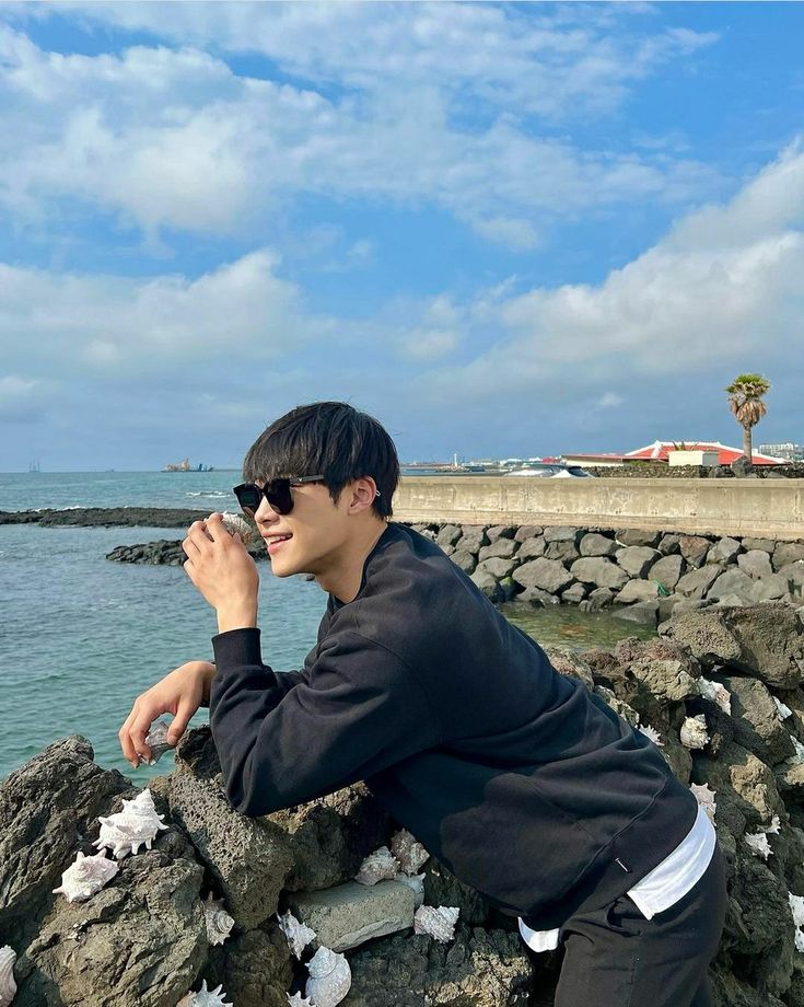 a young man sitting on rocks next to the ocean with his hand in his mouth