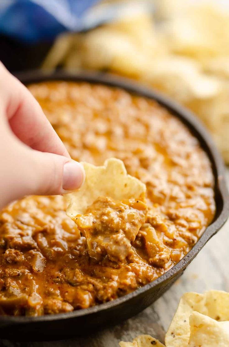 a hand dipping a tortilla chip into a skillet filled with chili and cheese