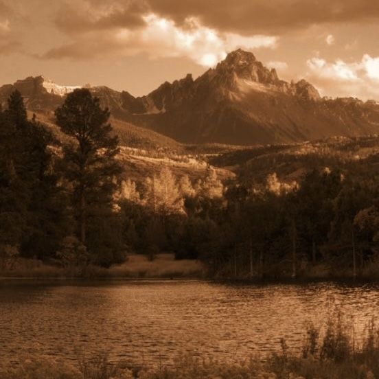 a lake surrounded by trees and mountains under a cloudy sky in sepia toned photograph