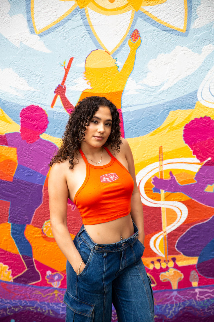 woman modeling upper park's club graphic baby rib halter top in orange in downtown chico, california standing in front of a mural. how to style halter top. cropped halter tank outfit. Stretch Cotton Halter Top For Day Out, Fitted Ribbed Halter Neck Crop Top, Casual Stretch Halter Top For Day Out, Stretch Halter Neck Casual Crop Top, Casual Stretch Halter Neck Crop Top, Casual Cropped Halter Top With Stretch, Casual Stretch Cropped Halter Top, Casual Cropped Stretch Halter Top, Spring Stretch Tank Halter Top